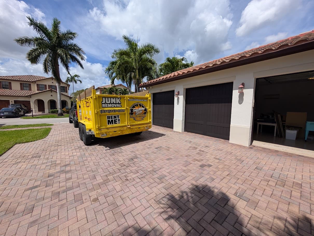Yellow dump trailer on a paver driveway beside a home with an open garage holding boxes and furniture