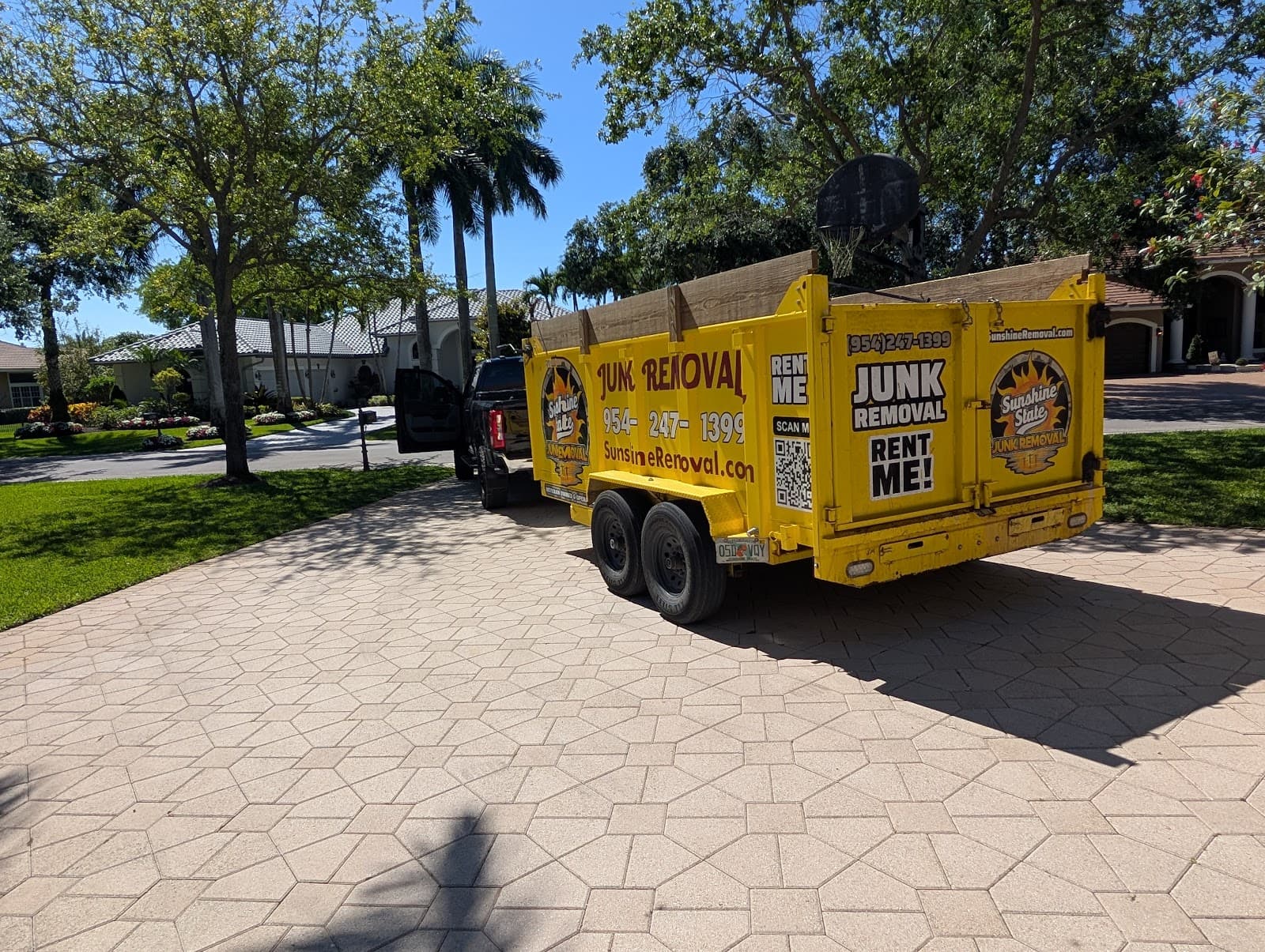 Yellow dump trailer hitched to a black pickup on a paver driveway in a residential neighborhood