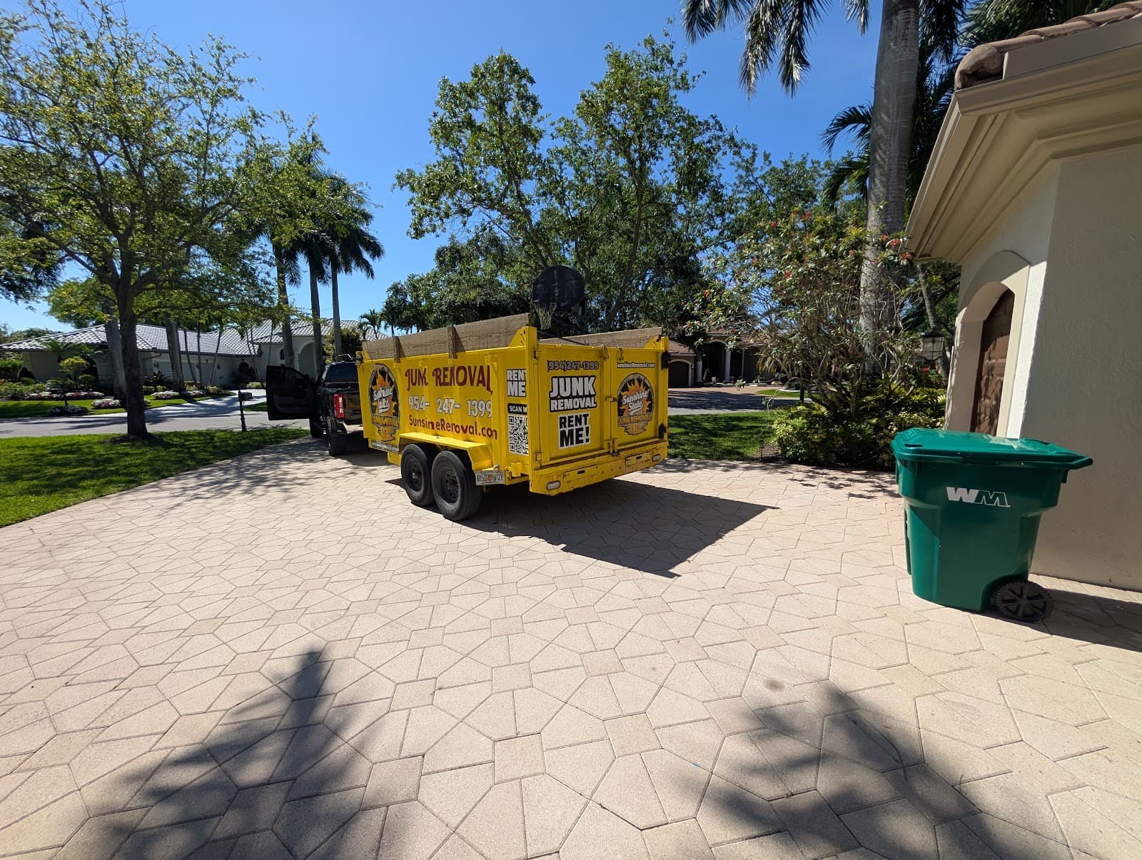 Yellow dump trailer and pickup parked on a wide paver driveway next to a green trash cart