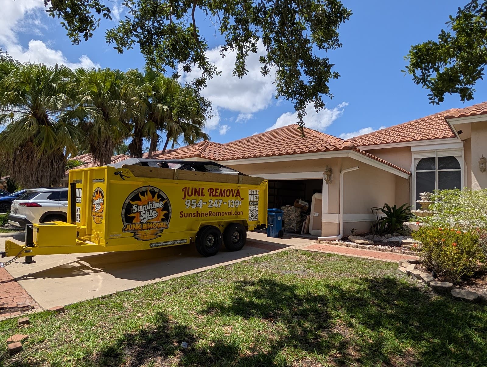 Loaded yellow dump trailer in the driveway of a tile-roof home with furniture and boxes in the open garage