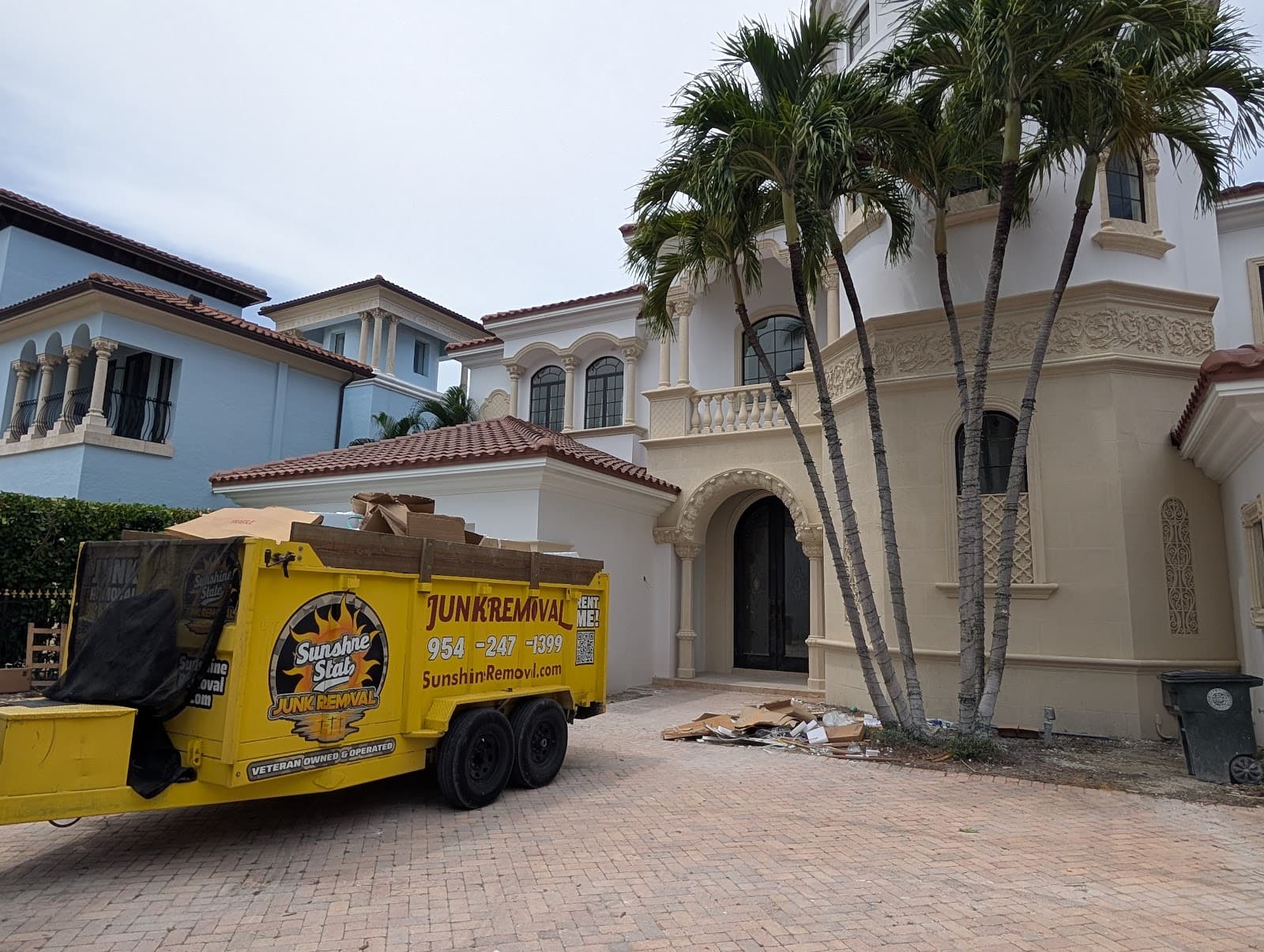 Yellow dump trailer loaded with cardboard outside a large white home, with debris piled near the entrance