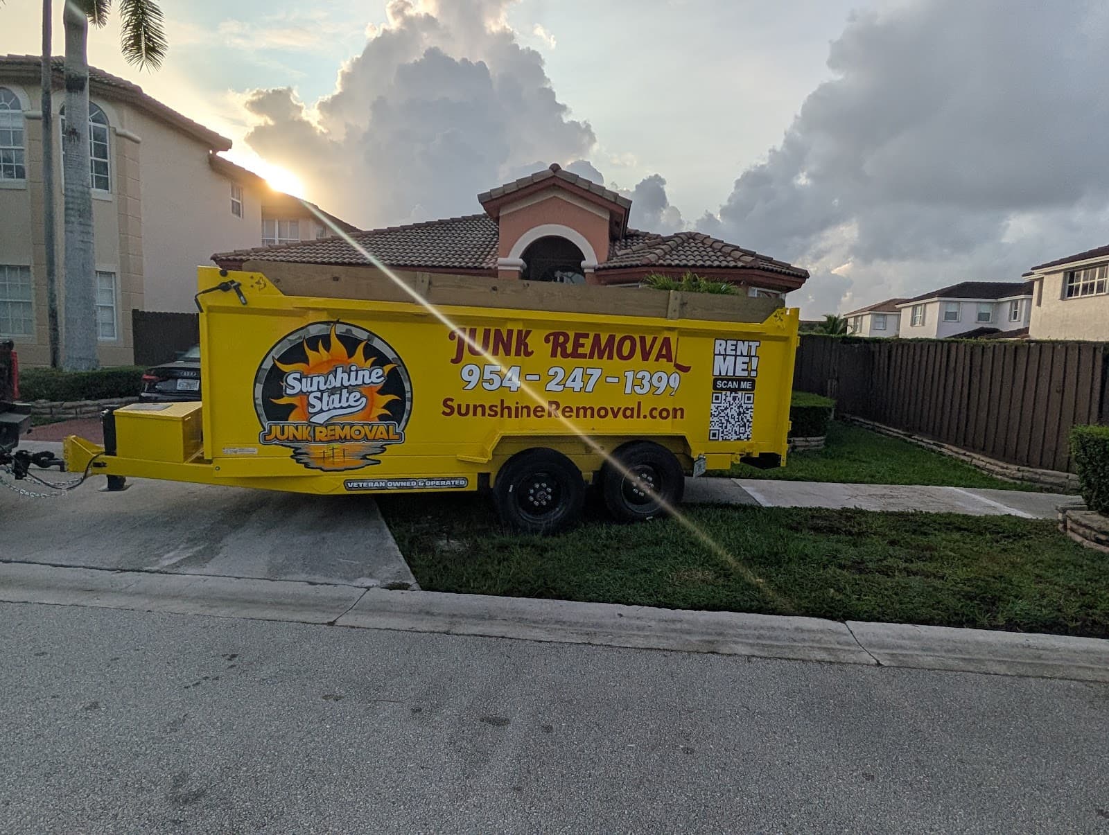 Yellow dump trailer parked at the curb in front of a two-story tile-roof home at sunset