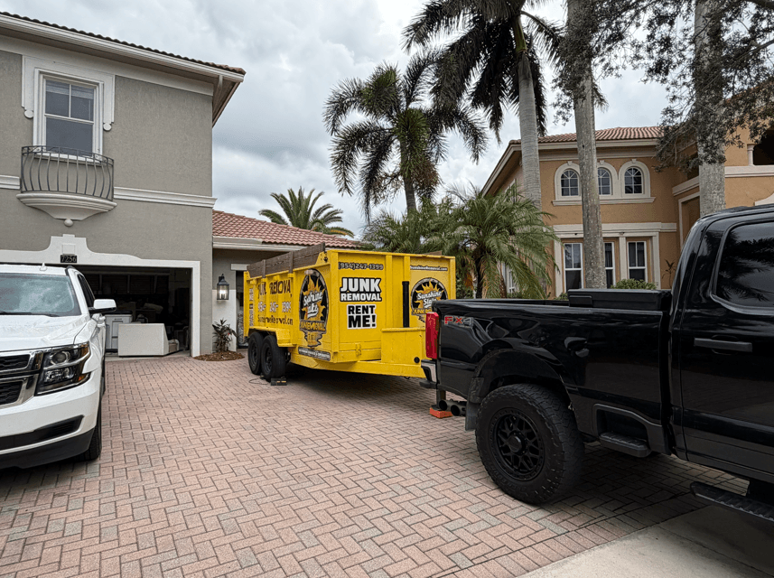 Yellow dump trailer on a paver driveway between a pickup and an SUV, with items in the open garage