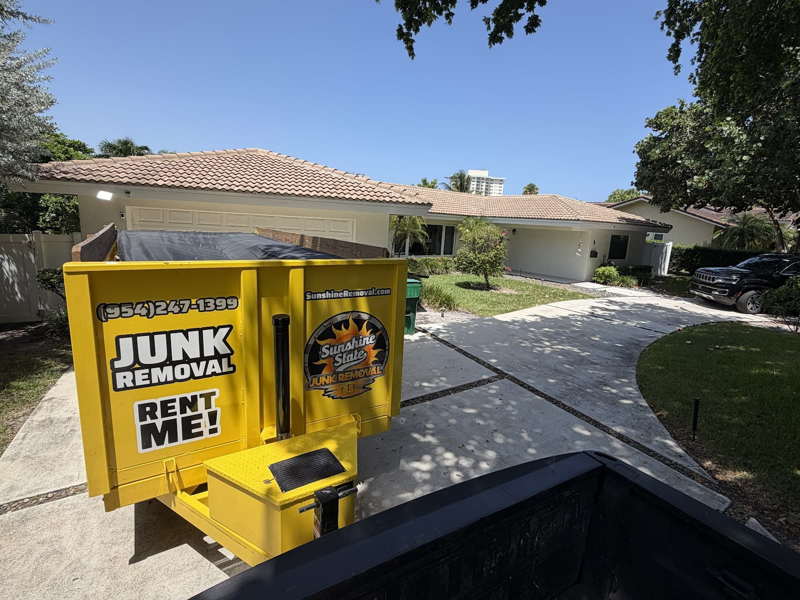 Tarped yellow dump trailer seen from a pickup bed in front of a single-story tile-roof home