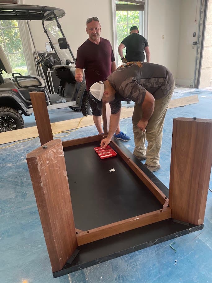 Three men around an upside-down wooden table on a floor covered in protective sheeting