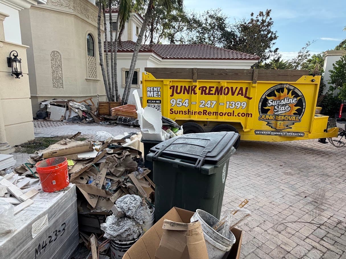 Yellow dump trailer on a paver driveway beside piles of cardboard, tile and buckets of construction debris