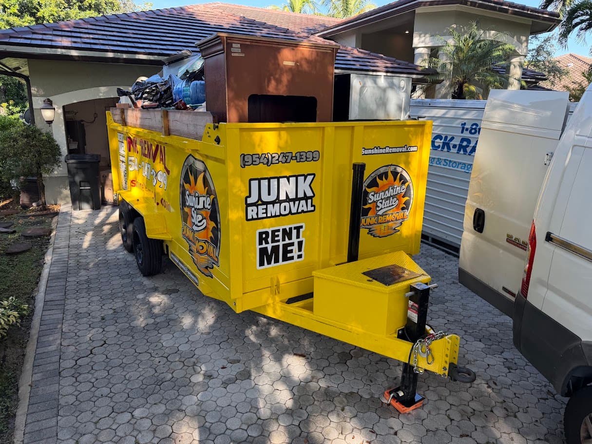 Yellow dump trailer piled with furniture and household items on a paver driveway next to a white van