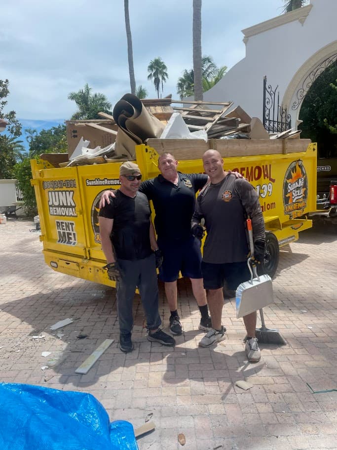 Three men with a broom and dustpan in front of a yellow dump trailer loaded with construction debris