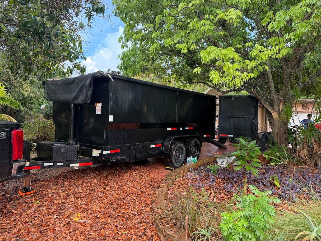 Black dump trailer with a tarp parked on a leaf-covered driveway under trees