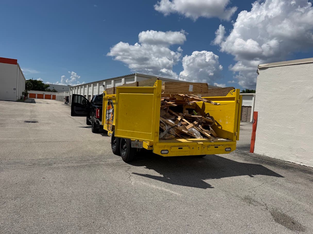 Yellow dump trailer loaded with broken wood pallets in a lot between rows of storage units