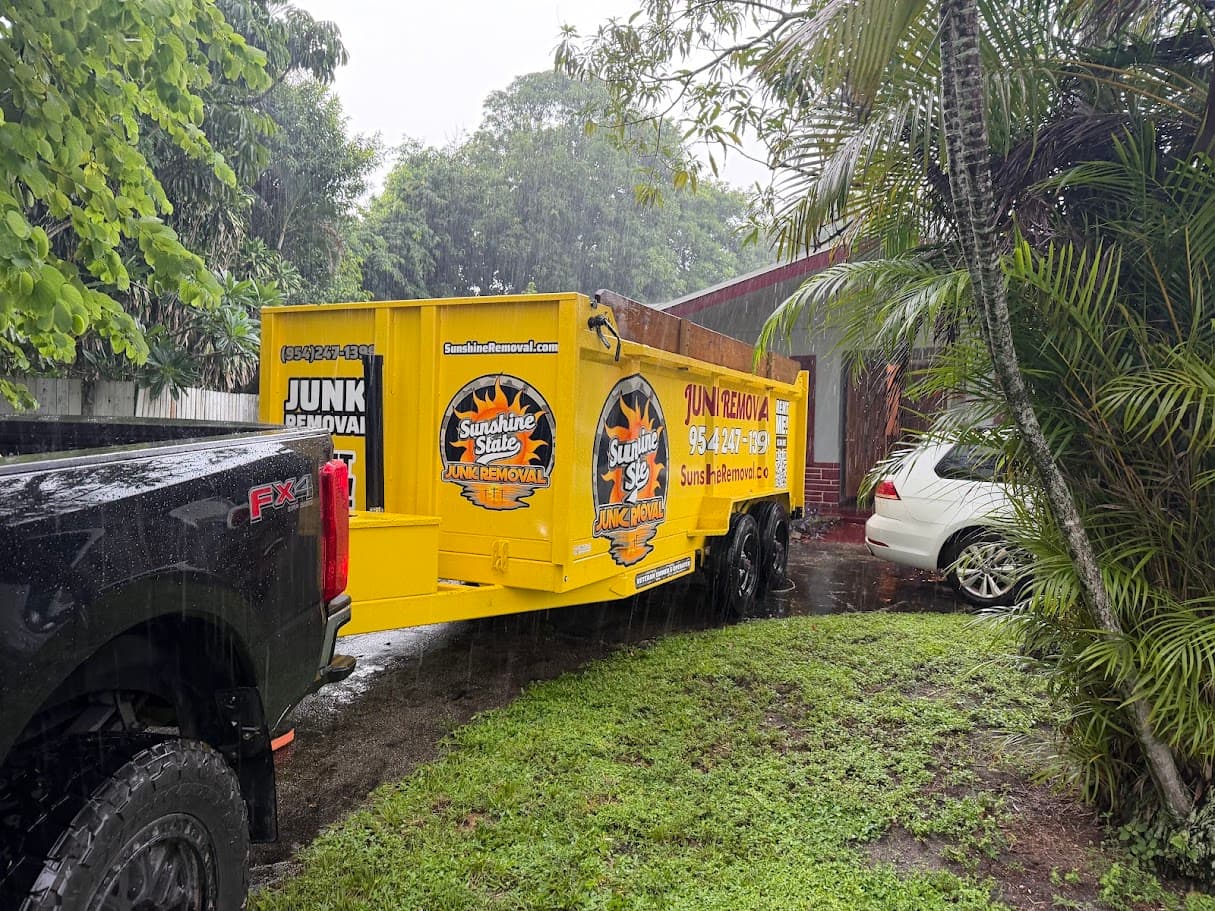 Yellow dump trailer hitched to a black pickup in the rain beside a house and palm trees